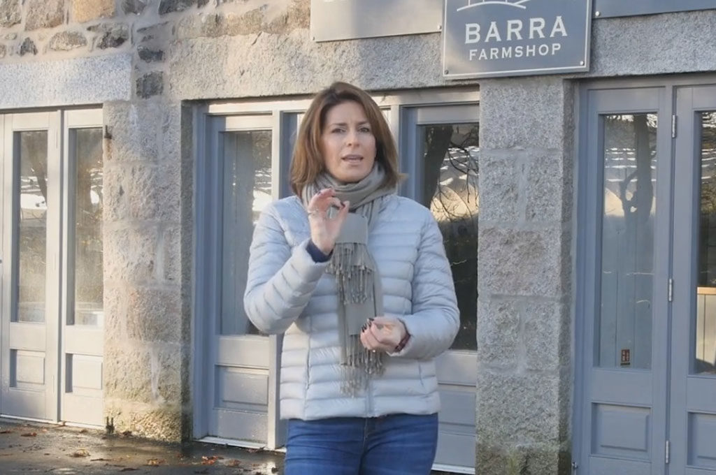 A person in a light jacket and scarf standing in front of "Barra Farmshop."