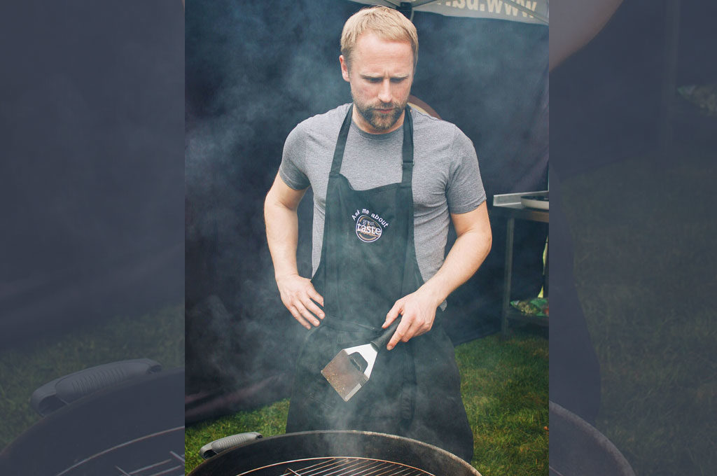 Craig Michie in an apron tending to a barbecue grill with smoke rising around.
