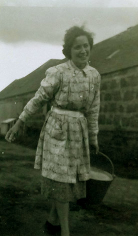 Person in a patterned dress holding a bucket, standing outside near a stone building.