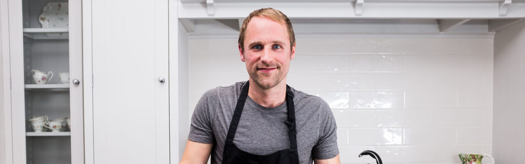 Person in a grey shirt and apron standing in a kitchen with white cabinets.